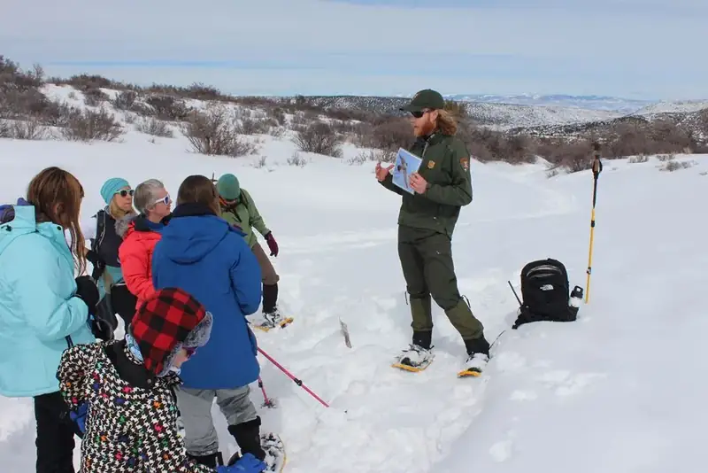 A park ranger in a green uniform holds up a photo to show a group of visitors standing in the snow