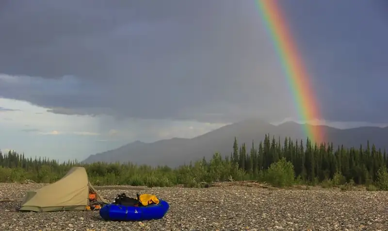 Green and Tan Tent with man's head peaking out of the flap, next to a blue packraft, situated on the stony riverbank. A rainbow appears in the sky in front of an incoming storm against mountains the background