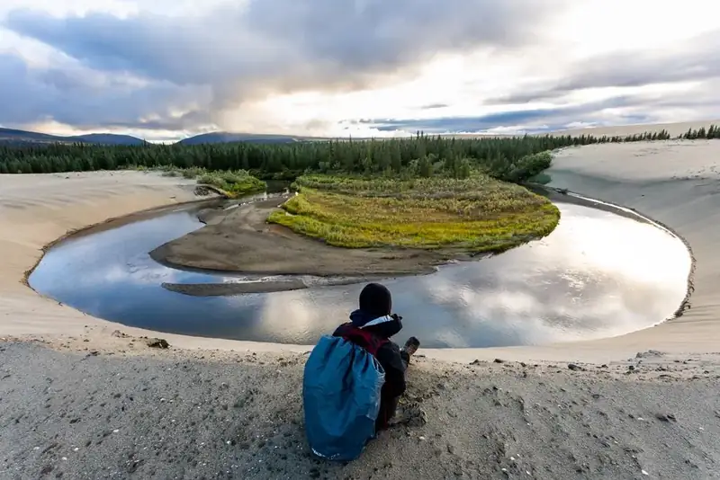Hiker stops near Niaktovik Creek. Sand dunes with creek and forest in background.