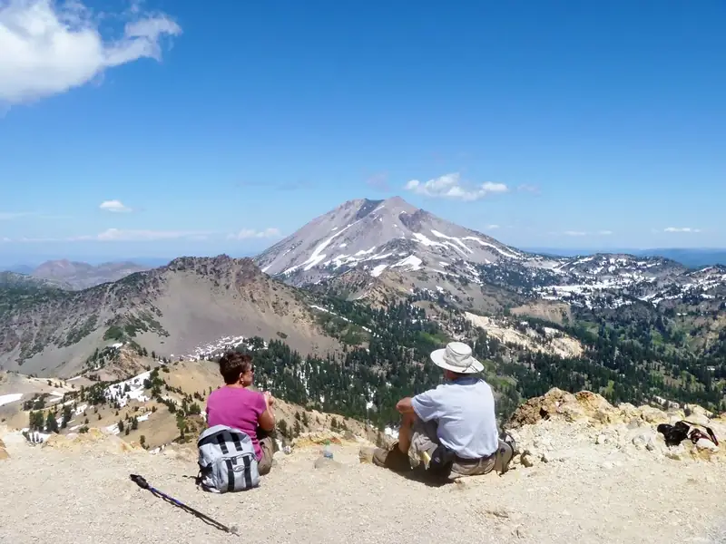 Two hikers sit on a mountain top with a view of multiple peaks, dotted with patches of snow 