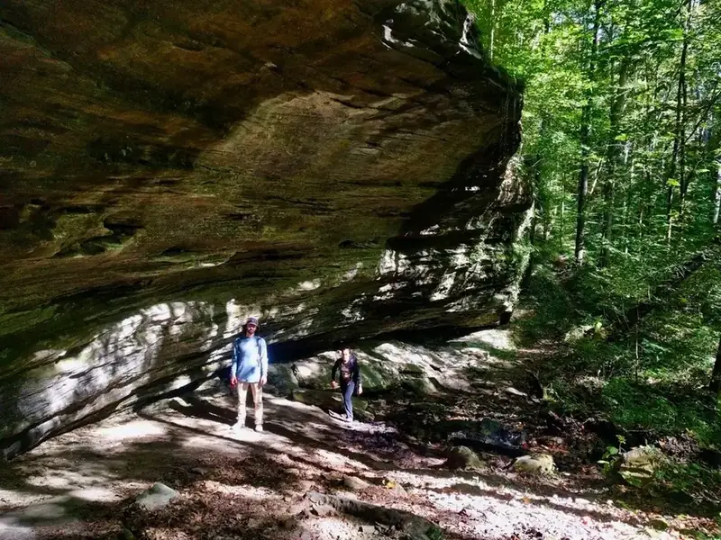 Two hikers stand next to a towering rock cliff with green foliage framing them.