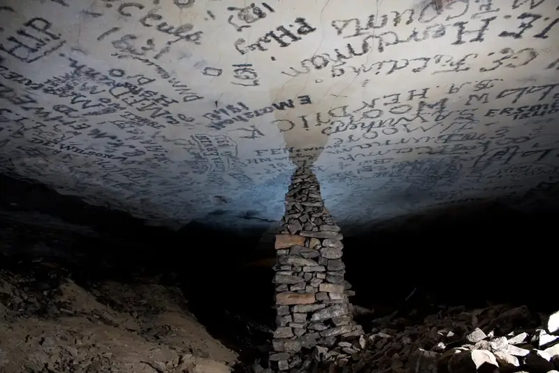 A large stacked stone pillar reaches the flat ceiling containing signatures in a cave passage. 