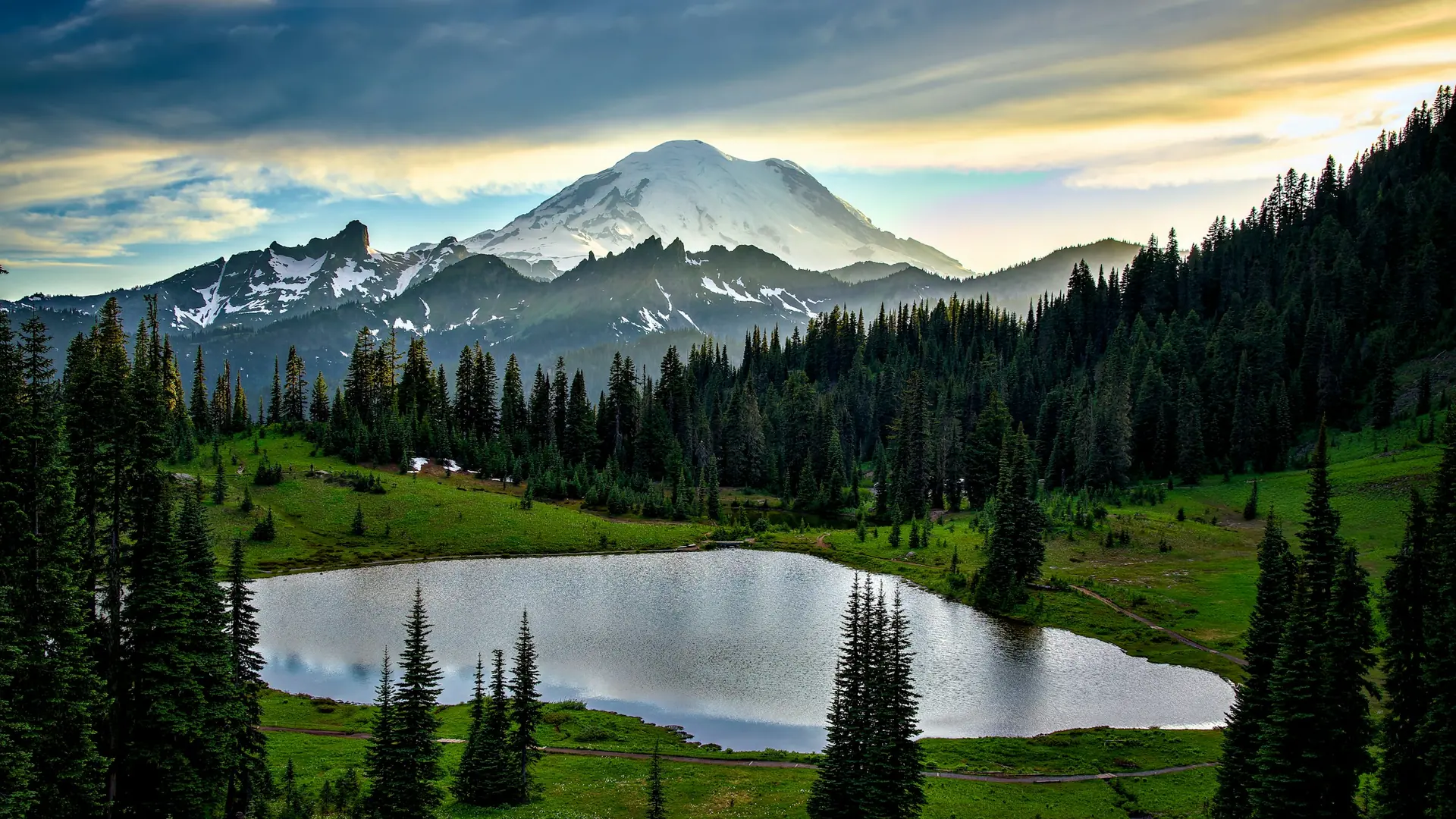 A small lake surrounded by evergreen trees below a snow-capped mountain peak