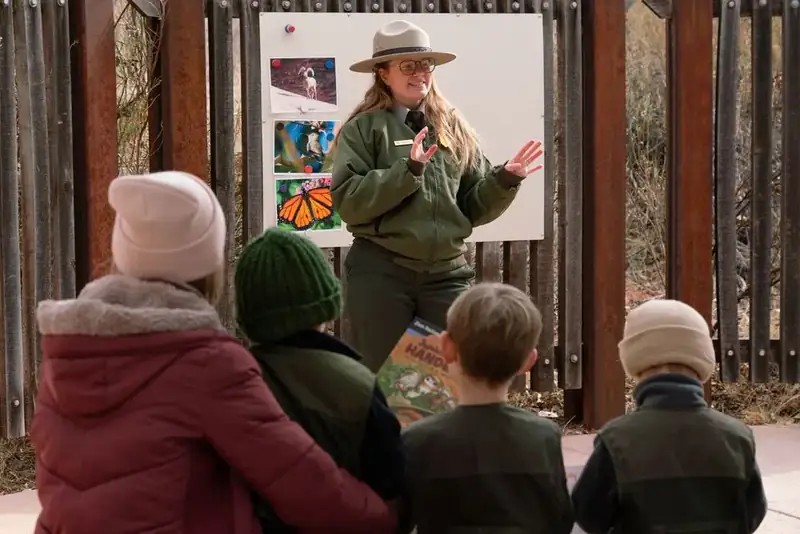 A ranger in a green coat stands in front of a seated family in hats and coats.